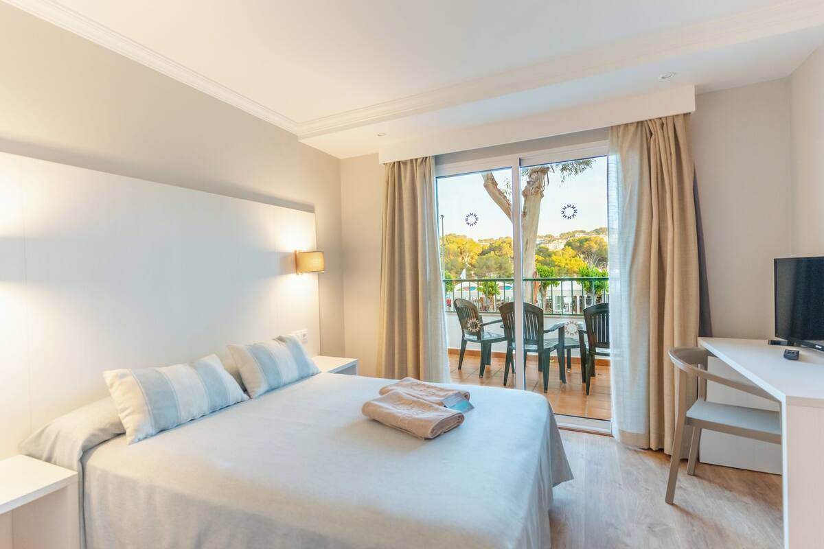 A hotel room with a bed, a desk, and a balcony with a view of trees and a clock.