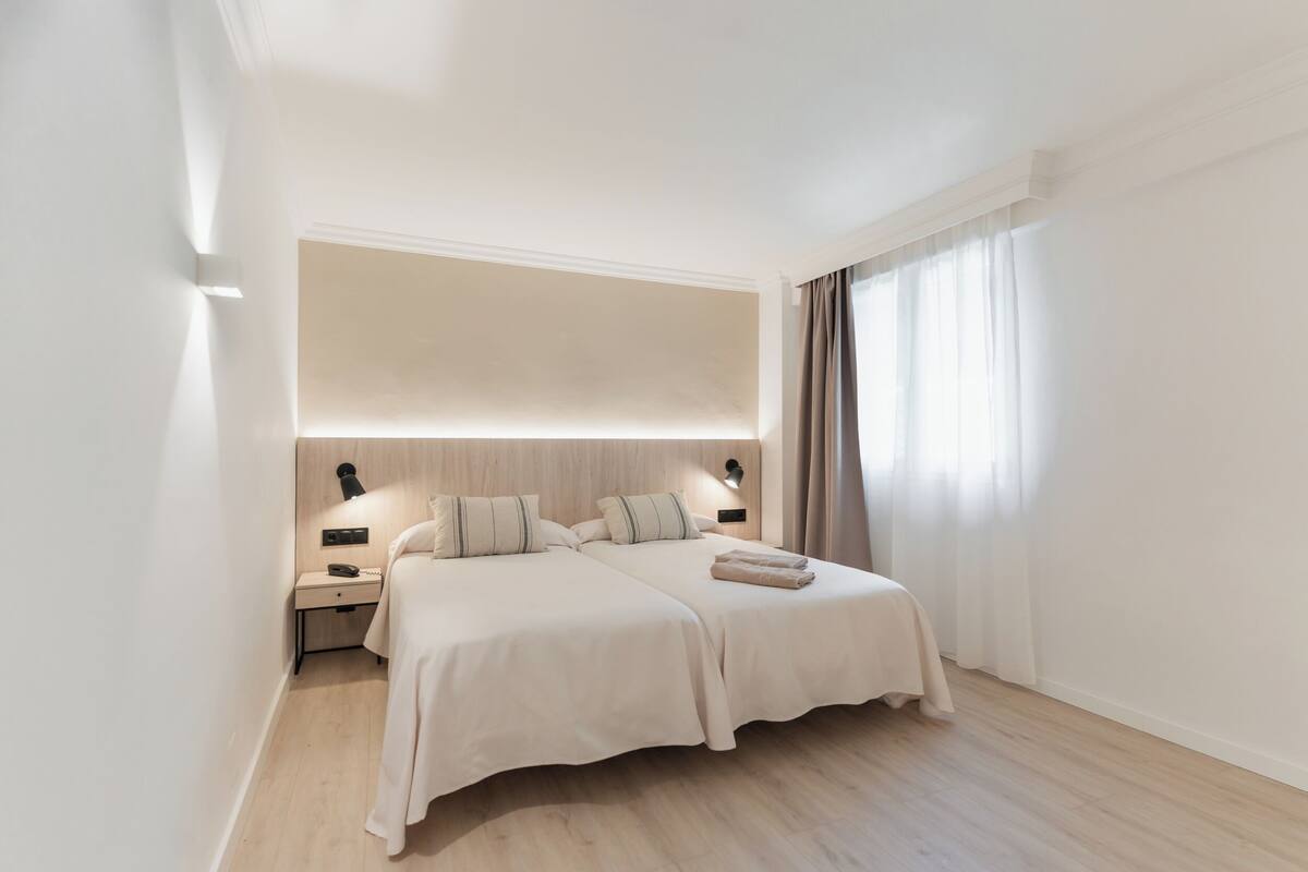 A modern hotel room with two beds, a bedside table with a lamp, and a large window with curtains.