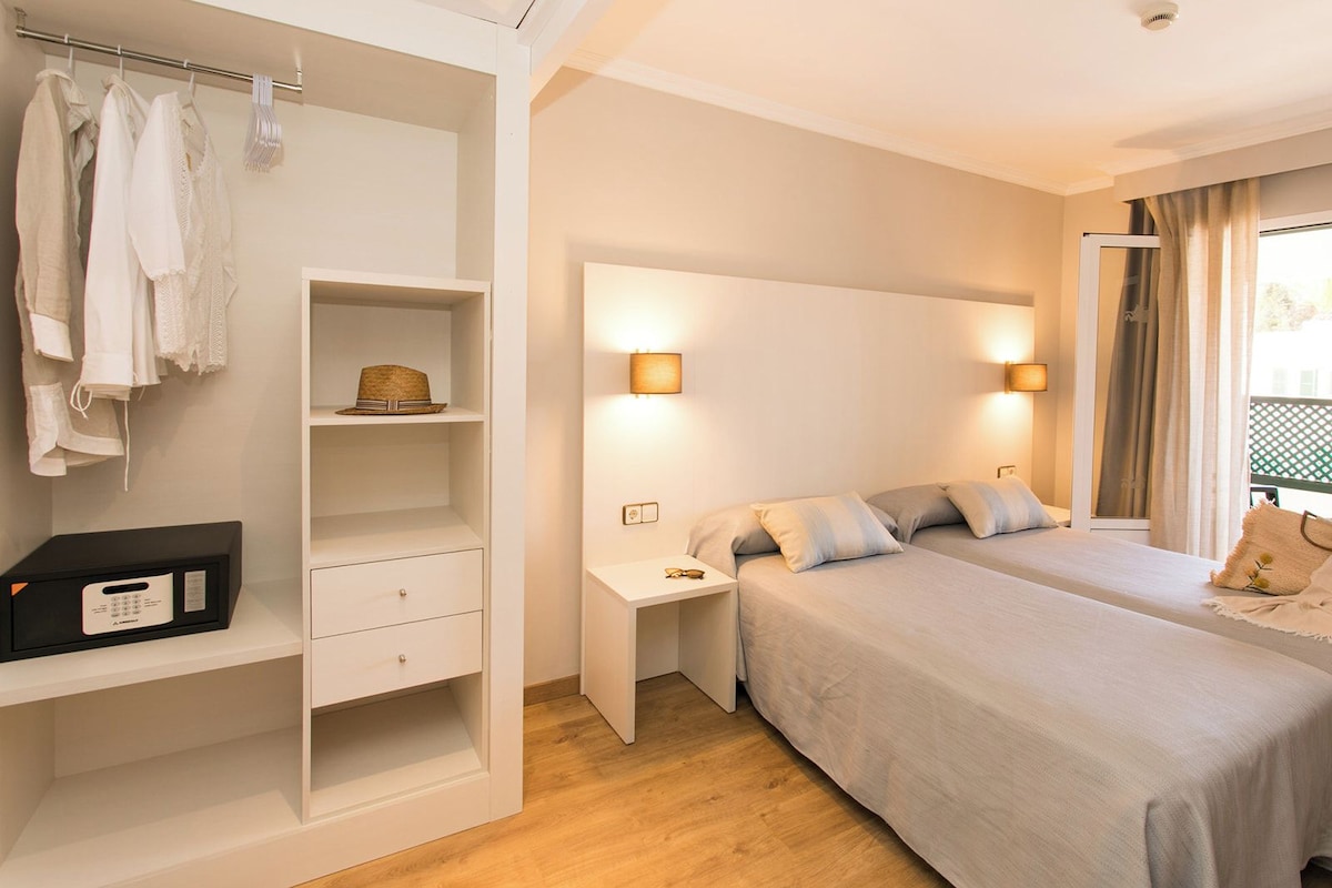 A hotel room with a wardrobe, two beds, bedside tables, and a window with curtains.