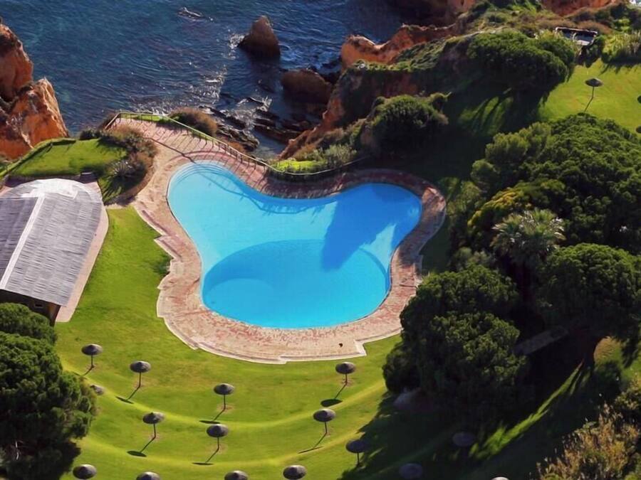 Vista aérea de una propiedad con una gran piscina, un edificio de techo inclinado y abundante vegetación.