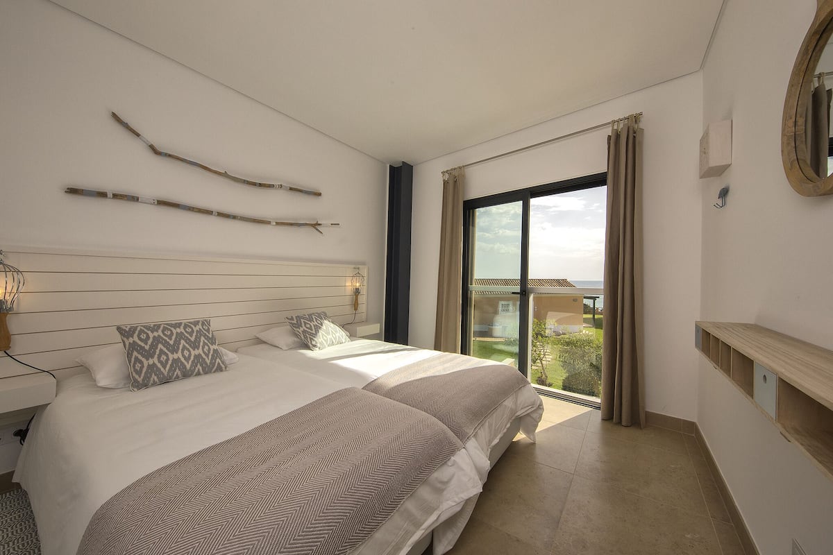 A bedroom with a large bed, a nightstand, a mirror, and a view of the outdoors.