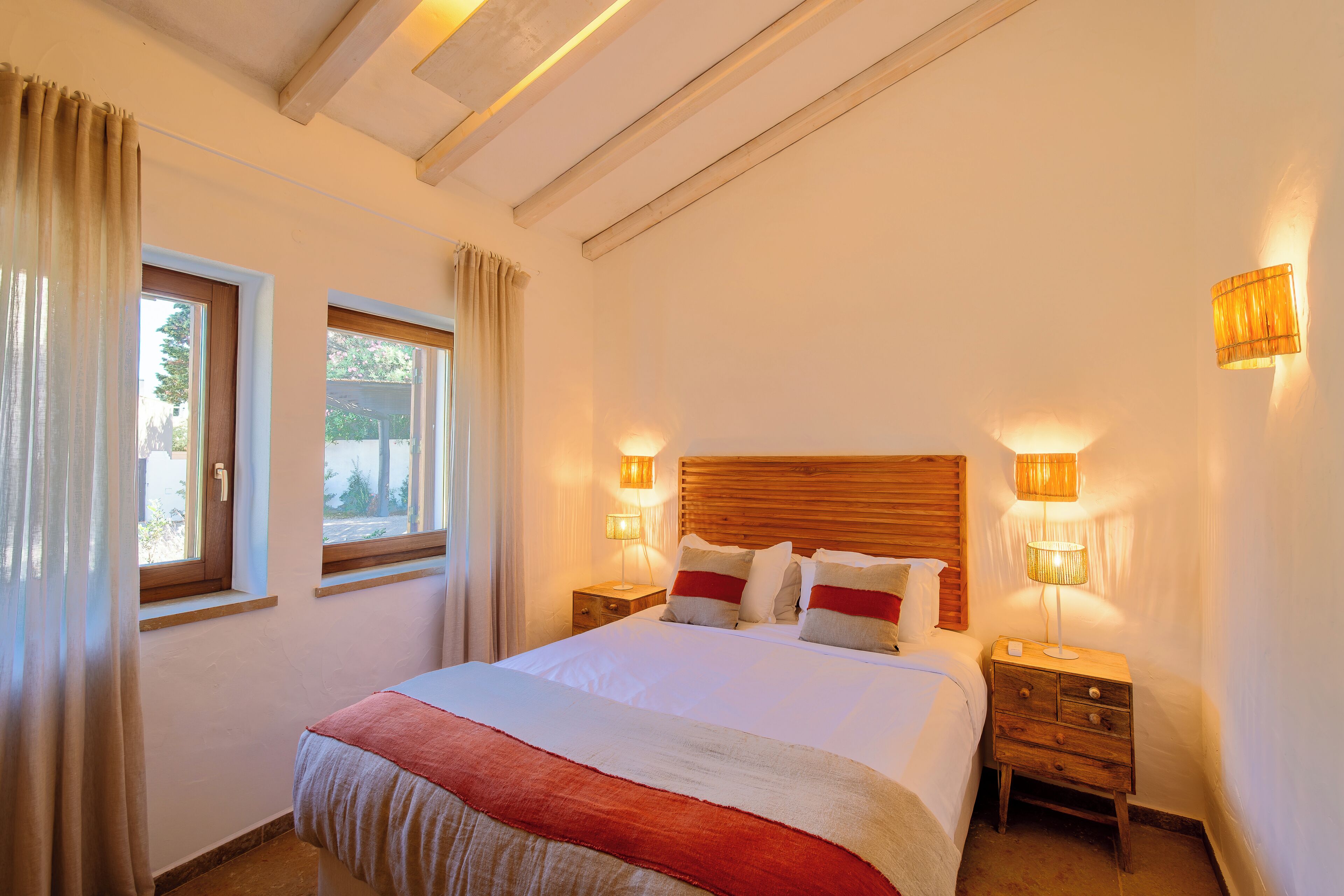 Ein Schlafzimmer mit einem großen Bett, zwei Nachttischen, einem Holzkopfende und zwei mit Vorhängen versehenen Fenstern.