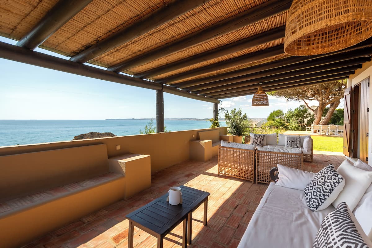 A covered outdoor seating area with a view of the sea, a sofa, a table, and wicker furniture.