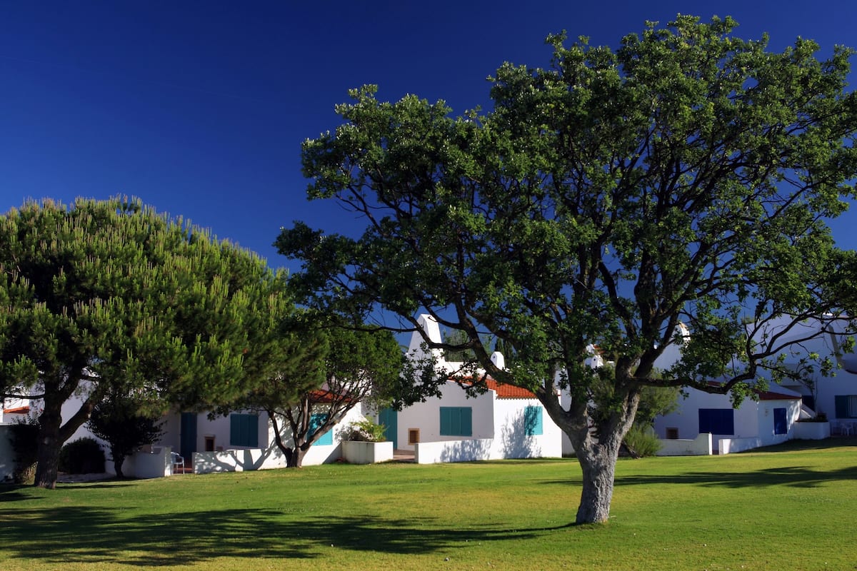 A white building with green shutters, surrounded by a grassy area and trees.