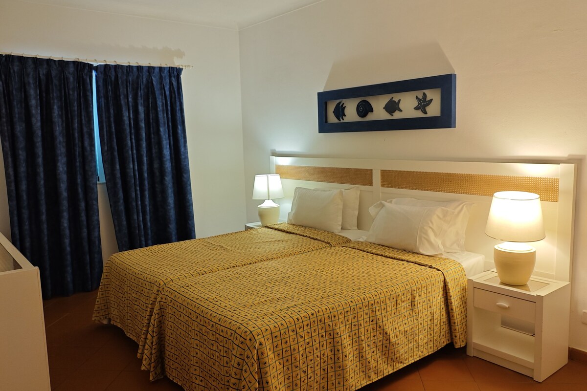 A hotel room with a bed, two bedside tables, a nightstand, a wall-mounted artwork, and a window with curtains.