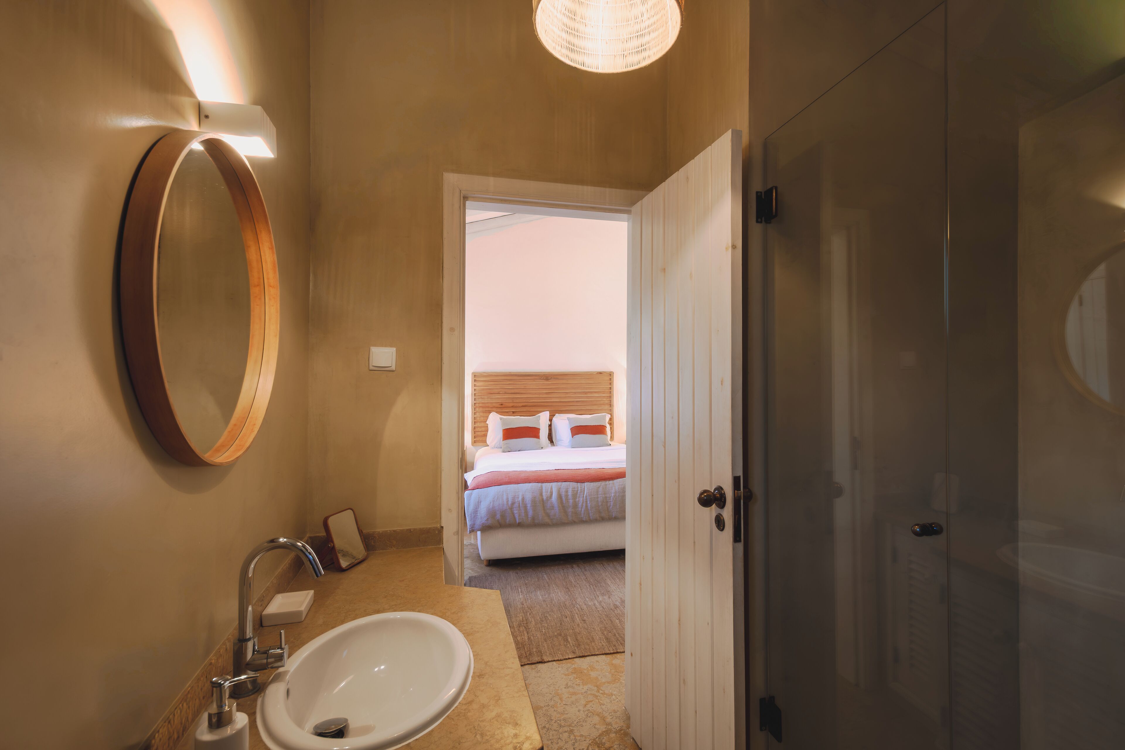 A bathroom with a sink, mirror, and a door leading to a bedroom with a bed and a window.