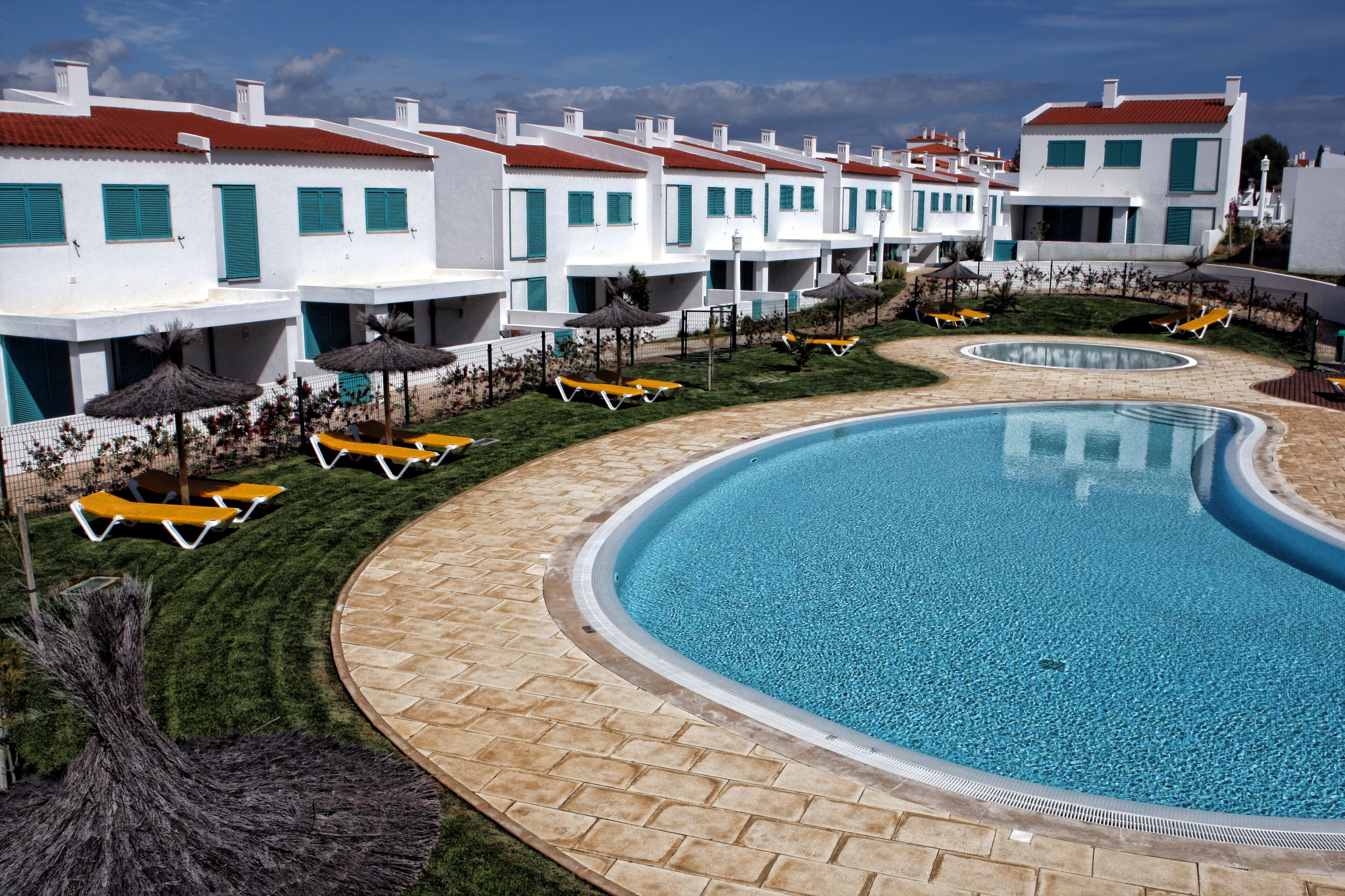 A resort with a large swimming pool, sun loungers, and white buildings with red roofs.