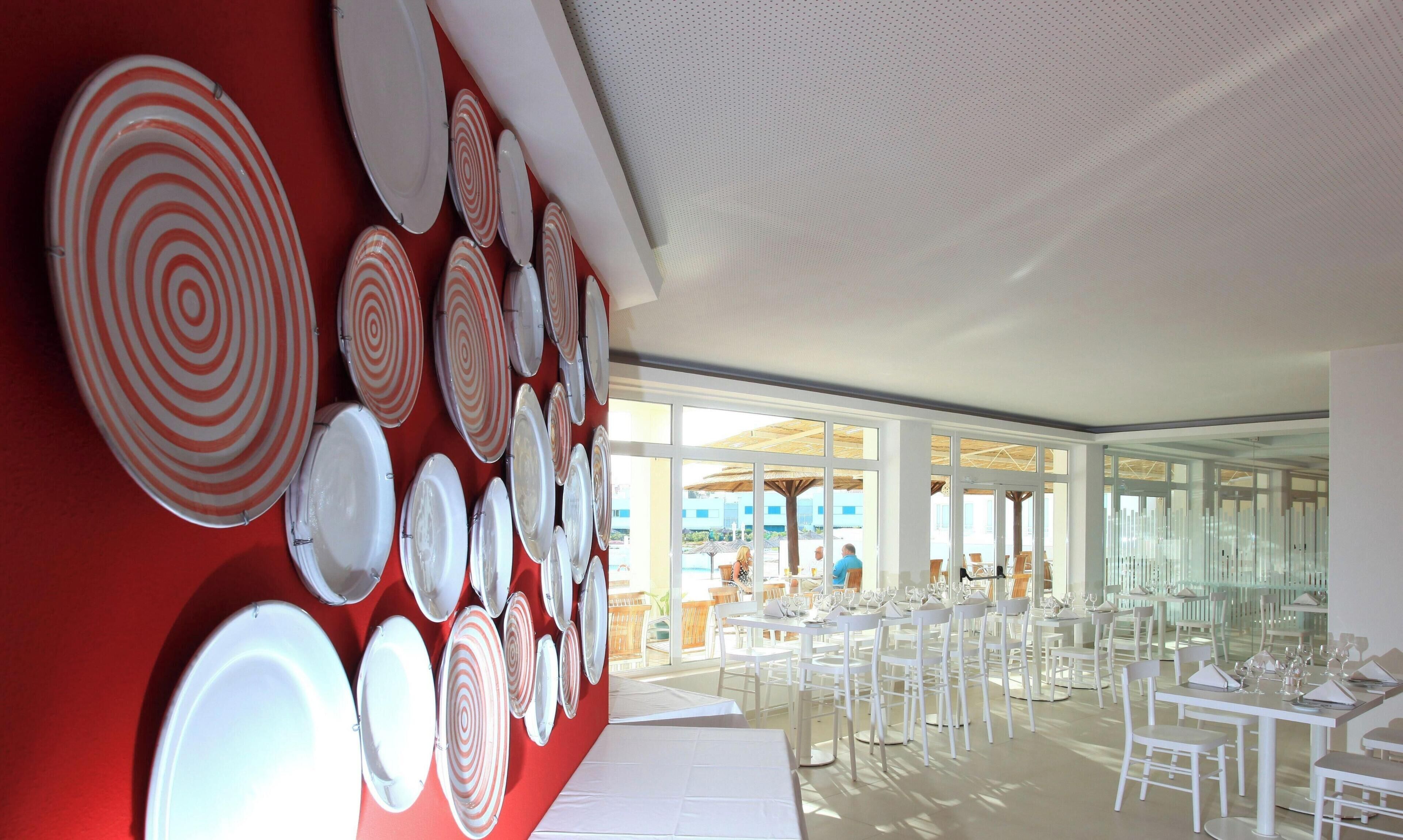 Un comedor moderno con mesas y sillas blancas, una pared roja con diseños circulares y ventanas amplias con vistas al exterior.