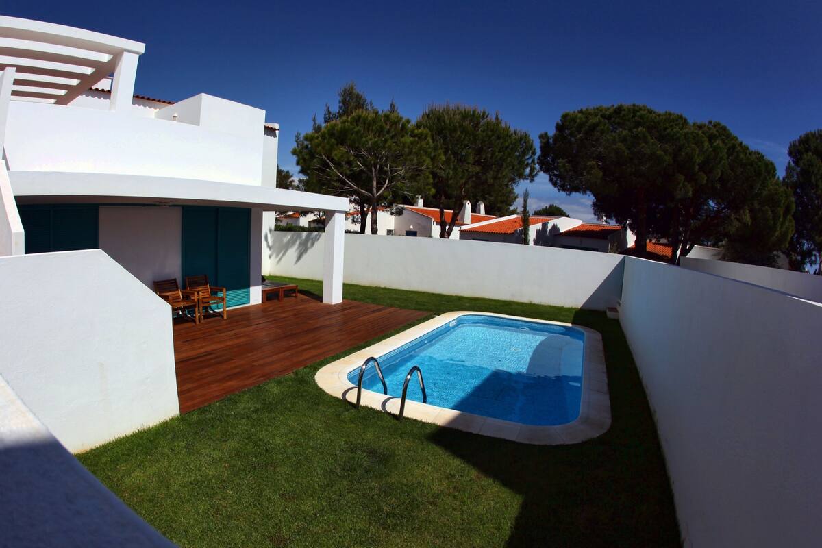 A modern outdoor pool area with a wooden deck and seating, surrounded by white walls and a clear blue sky.