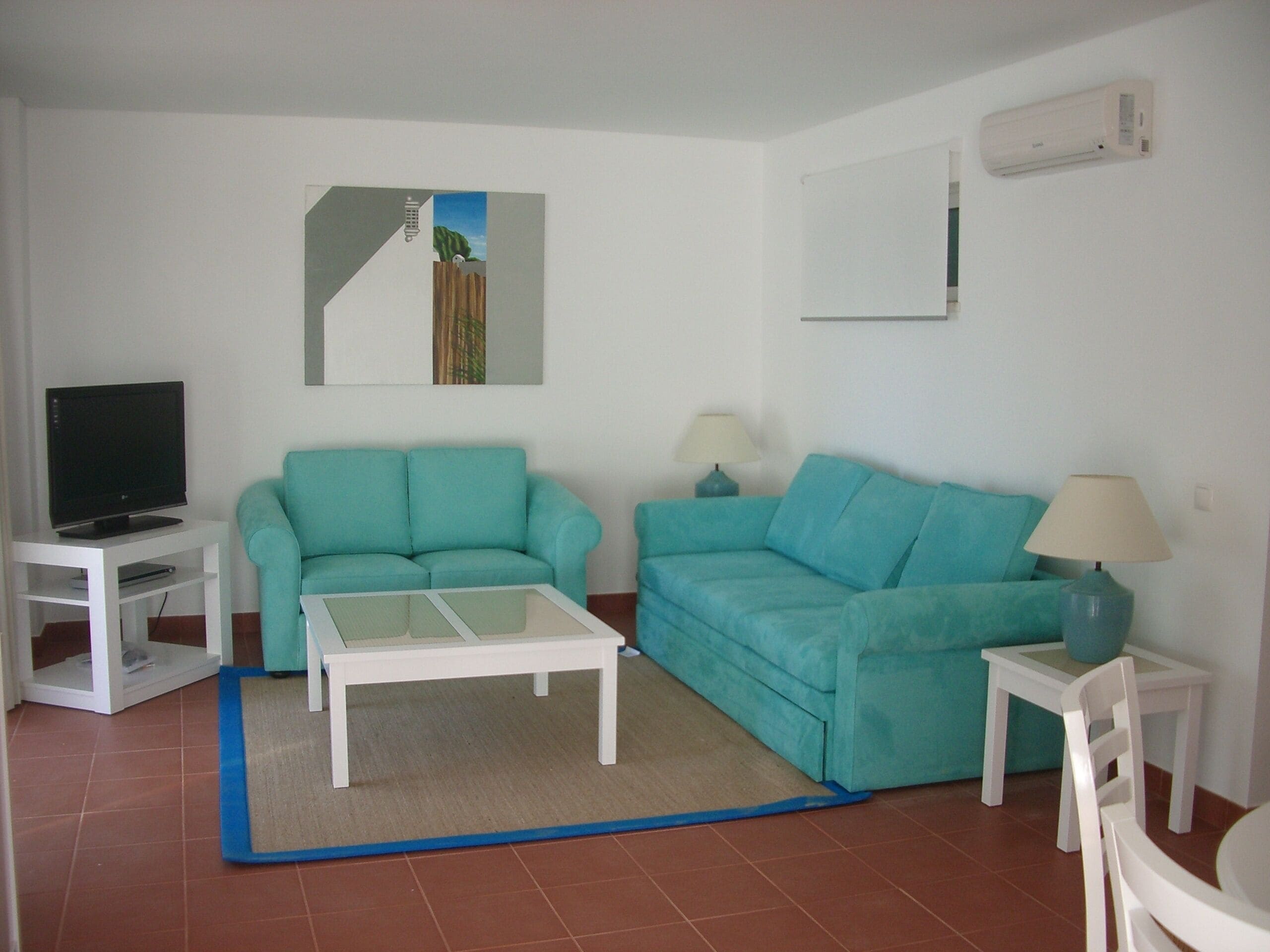 Una sala de estar con dos sofás color turquesa, una mesa de centro blanca, un televisor sobre un soporte blanco y una mesita con una lámpara.