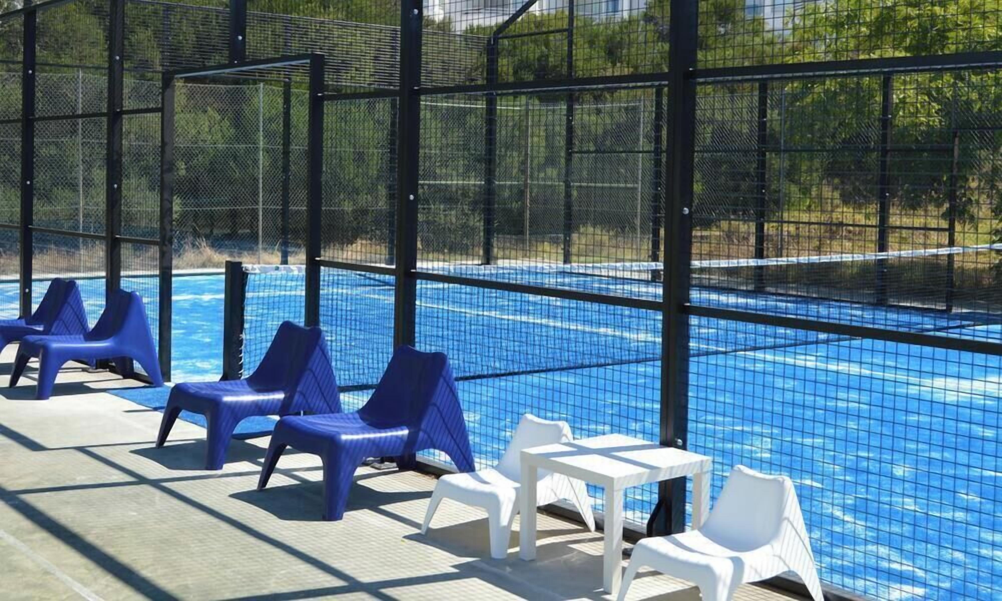 A swimming pool with blue water, surrounded by a black fence. There are blue and white lounge chairs and a small white table.