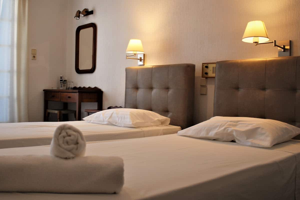 Habitación de hotel con dos camas, cada una con cabecera y lámpara de noche. Hay una mesita de noche con espejo y lámpara, y una ventana con cortinas.