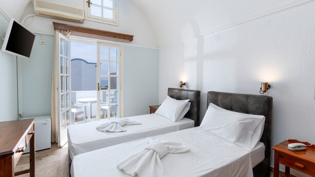 A hotel room with two beds, a TV, a desk, and a balcony with chairs. - Artemis Hotel (Santorini)