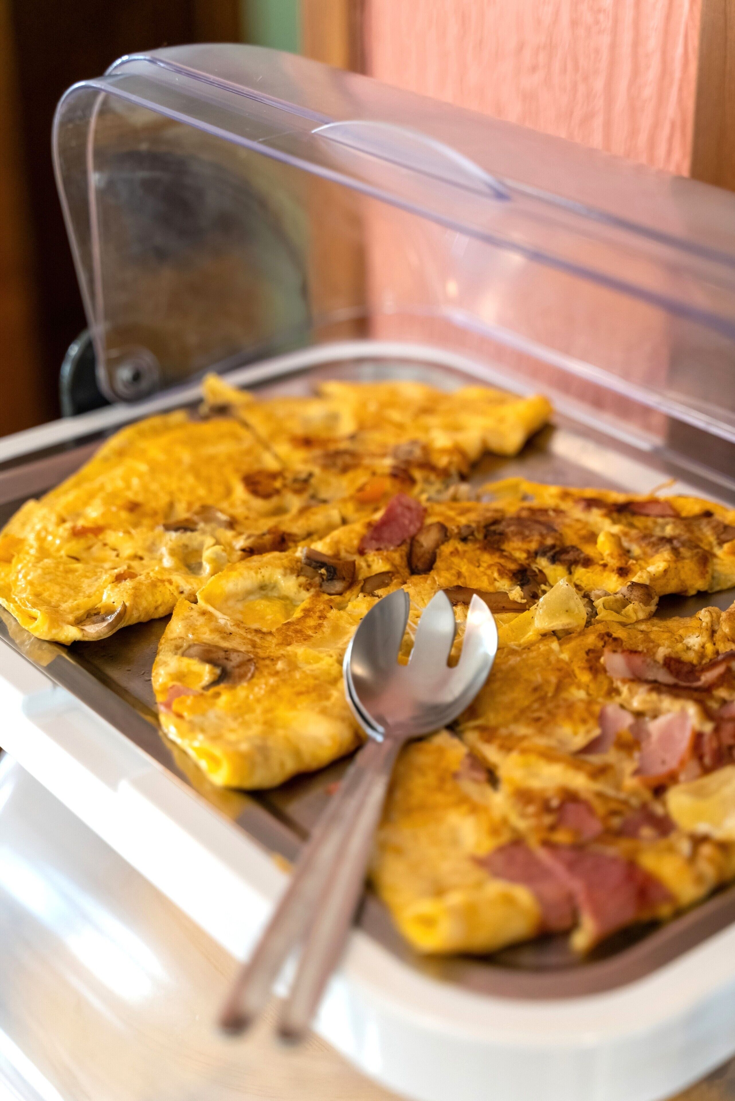 A breakfast tray with a slice of omelette and ham, a spoon, and a plastic cover.