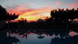 A swimming pool reflecting a sunset sky with silhouettes of trees and a mountain in the background. - Artemis Hotel (Santorini)
