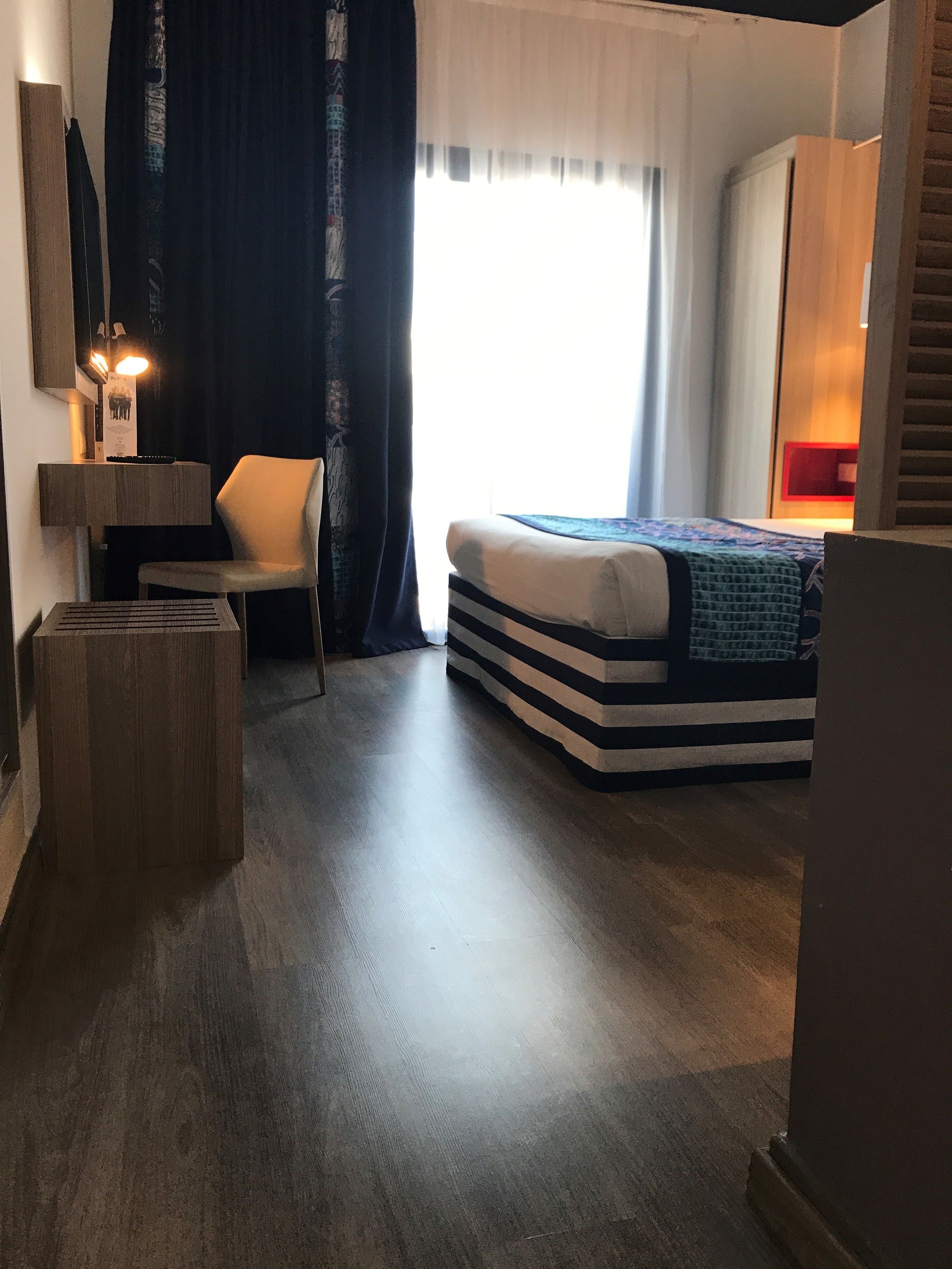 Una habitación de hotel moderna con una cama, una silla, un escritorio y un ventanal con cortinas.