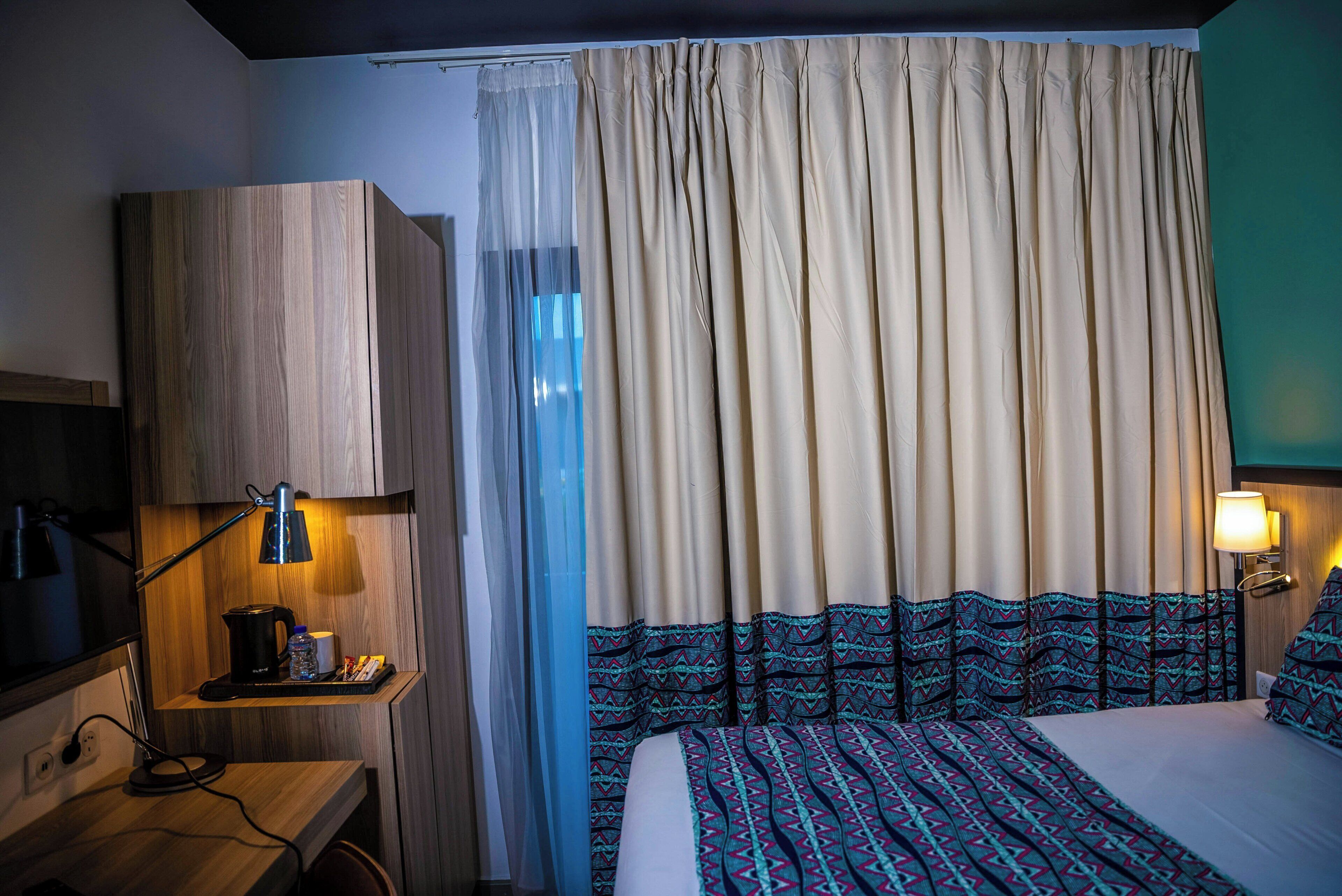 Una habitación de hotel con cama, escritorio con lámpara, cafetera y cortinas.