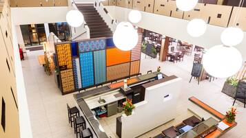A modern hotel lobby with a central reception desk, seating areas, and a staircase leading to upper floors.