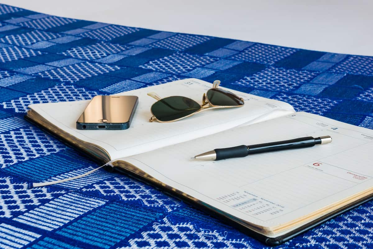 Un lit bien fait, recouvert d’une couvre-lit à motifs bleus, un smartphone, des lunettes de soleil et un carnet avec un stylo.