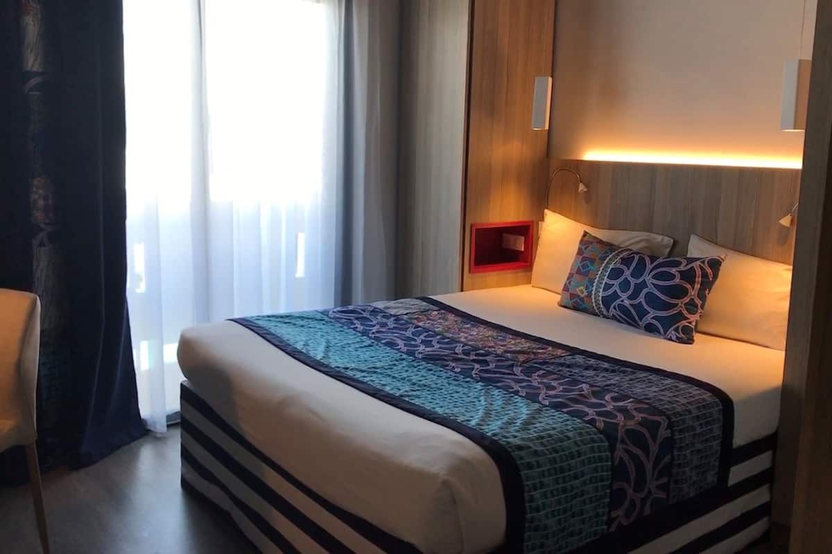 Une chambre d’hôtel avec un grand lit, une tête de lit en bois, une table de chevet avec un détail rouge et une fenêtre ornée de rideaux légers.