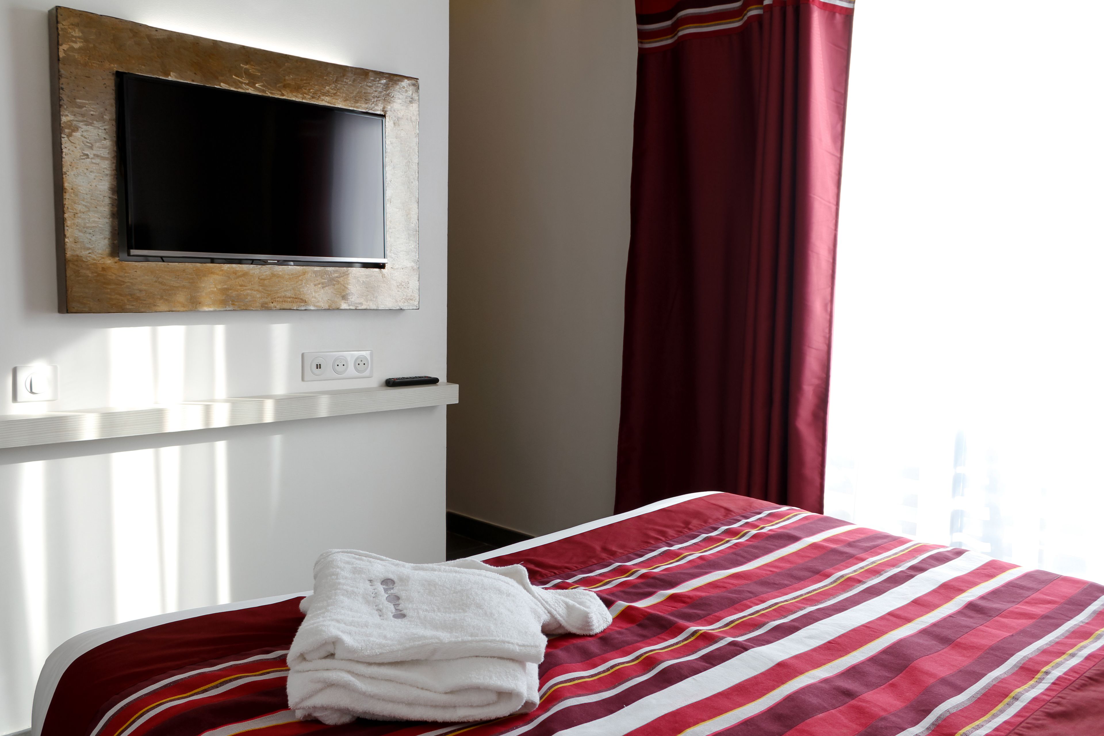 Una habitación de hotel con cama, televisor montado en la pared, enchufe y ventana con cortinas.