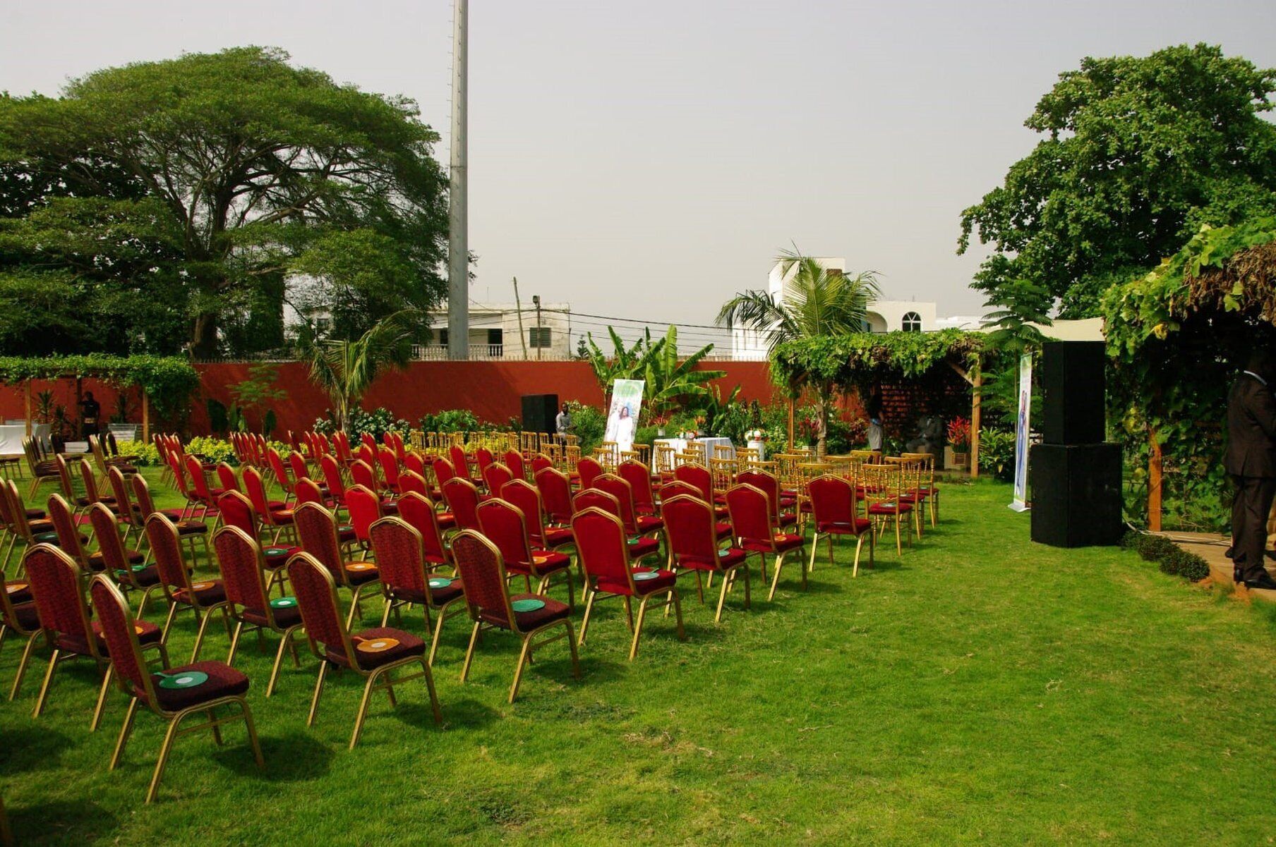 An outdoor seating area with rows of red chairs and tables, surrounded by greenery and a building in the background.