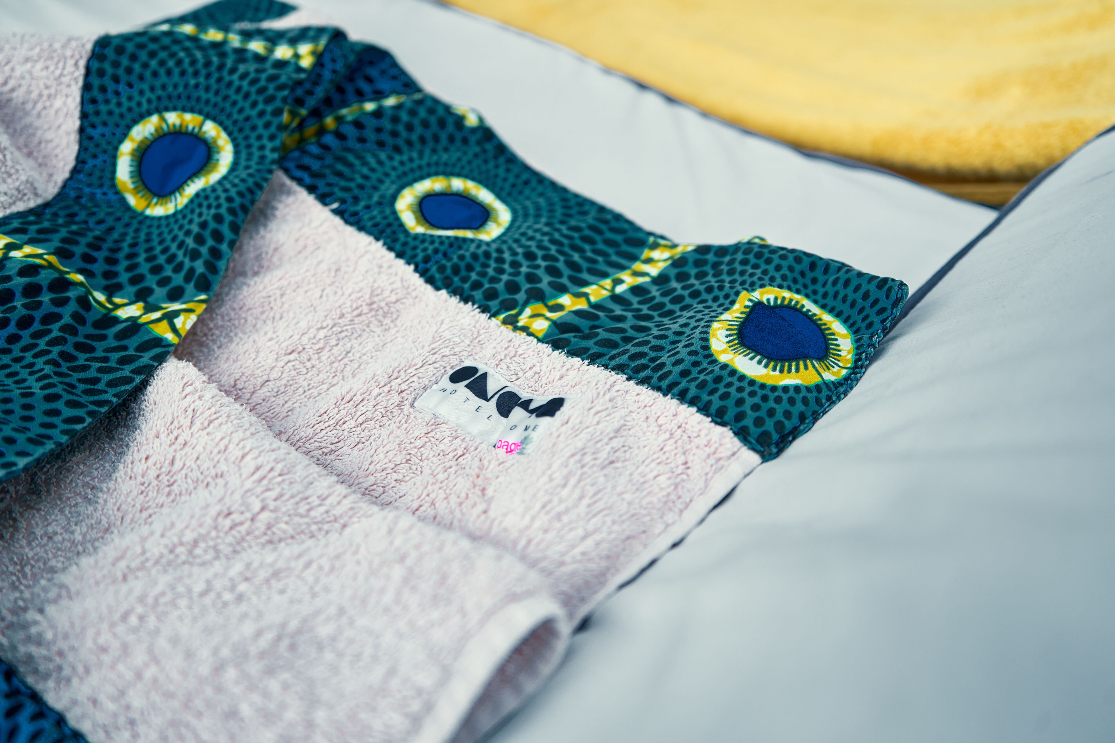 A bed with a white sheet and a green and blue patterned blanket with a small embroidered logo.