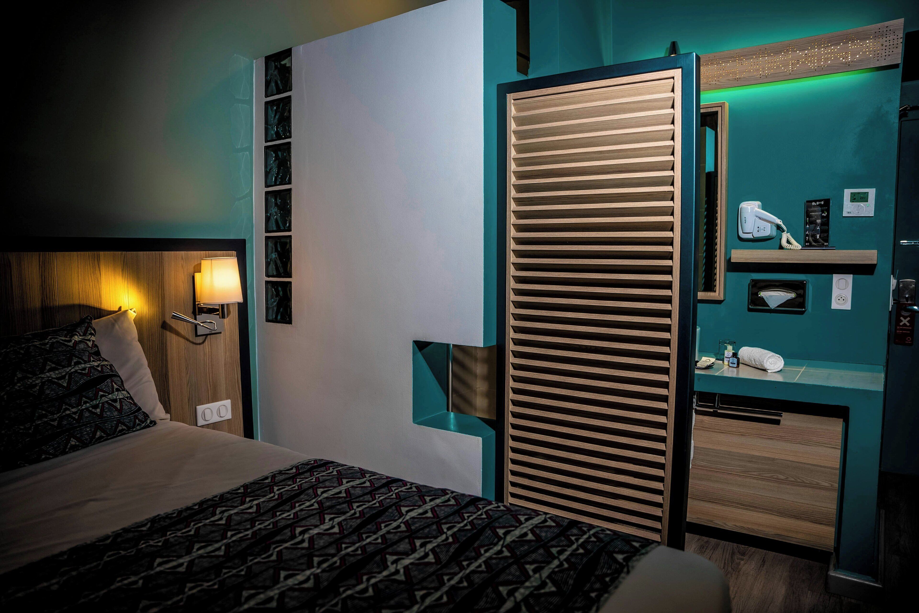 Habitación de hotel con cama, mesita de noche, lámpara, espejo, estante con artículos de aseo y un armario de madera.