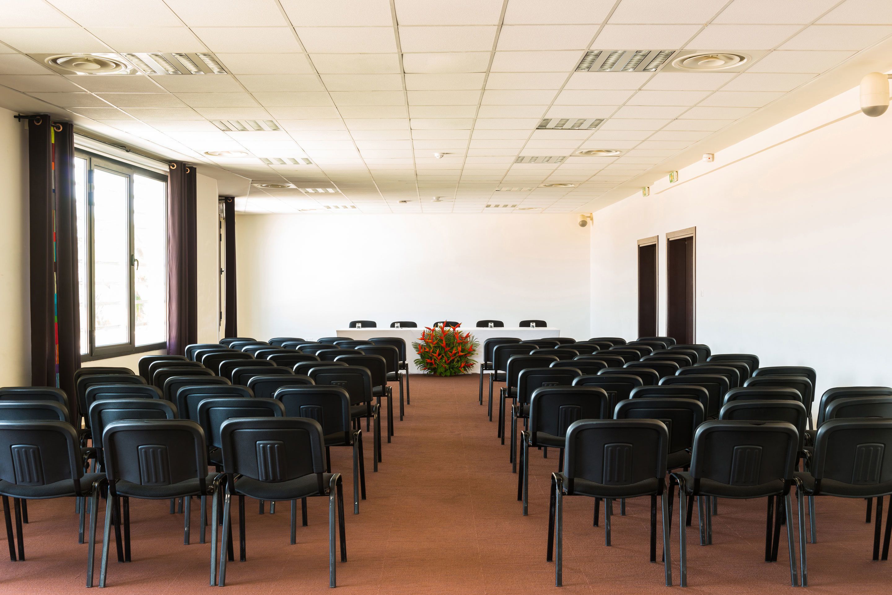 Une salle de conférence avec des rangées de chaises noires disposées face à une table blanche agrémentée d’un arrangement floral.