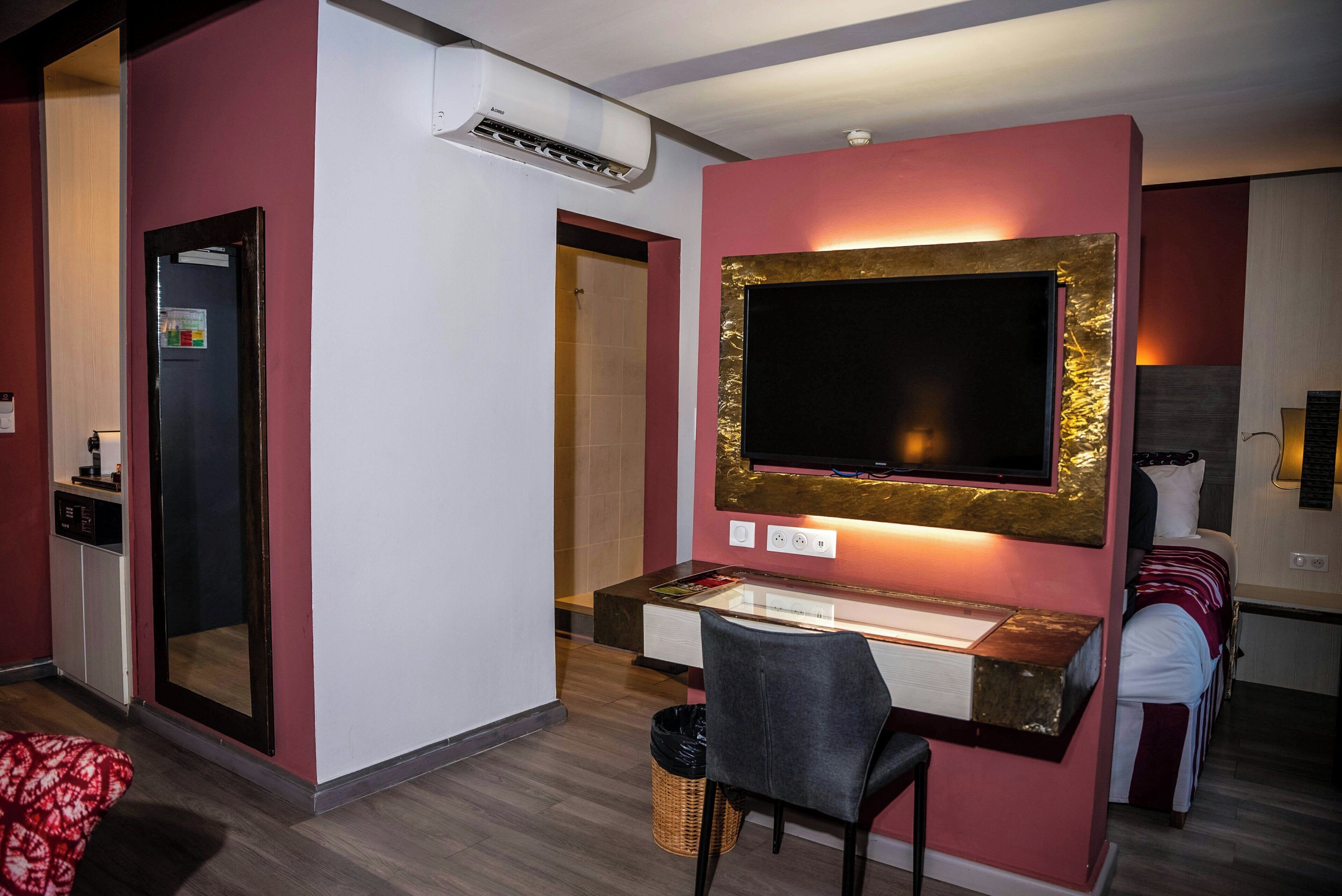 A hotel room with a bed, a desk with a chair, a TV, a mirror, an air conditioning unit, and a small trash bin.
