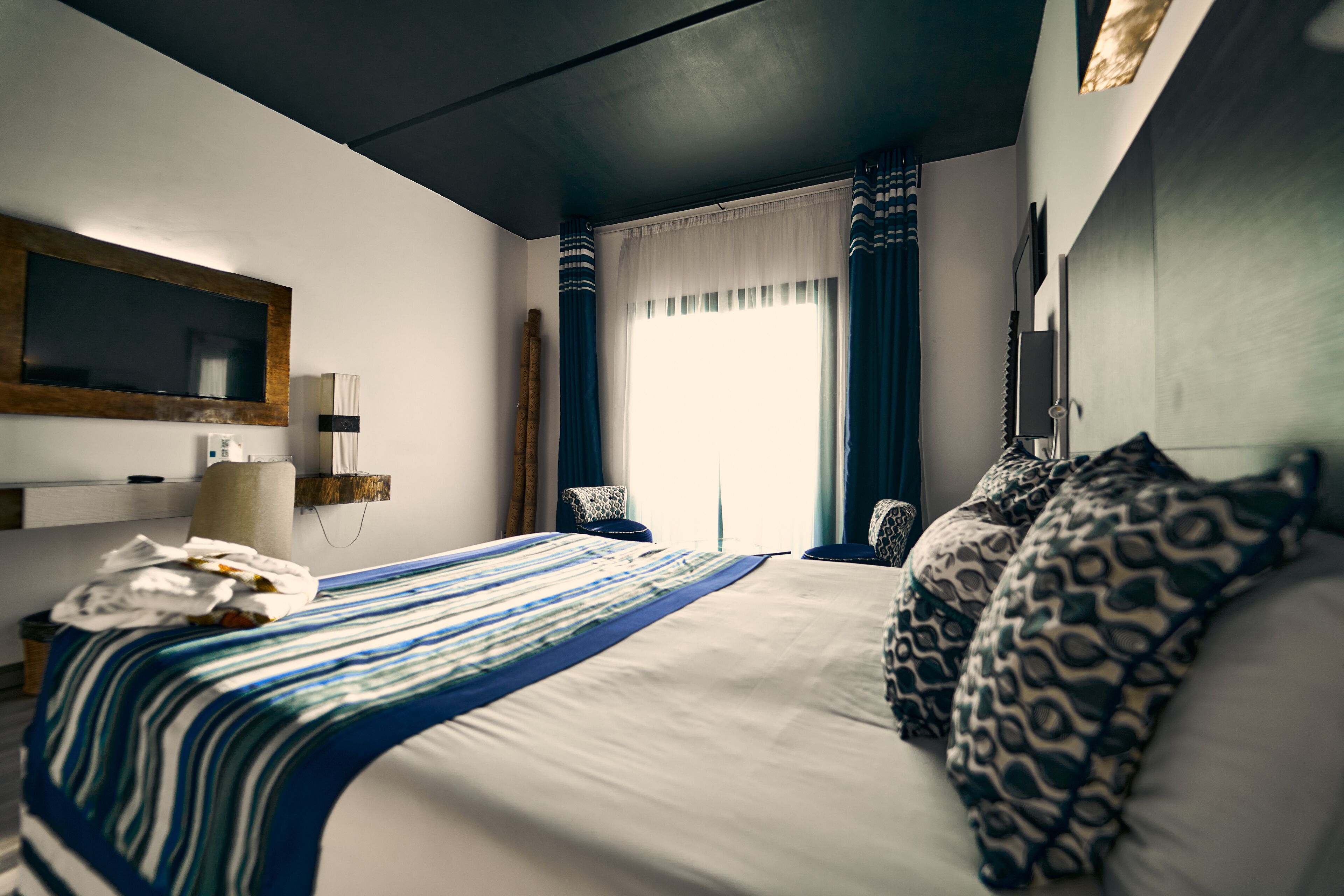 Una habitación de hotel moderna con una cama grande, un televisor de pantalla plana en la pared, una mesita de noche con una lámpara y una ventana con cortinas.