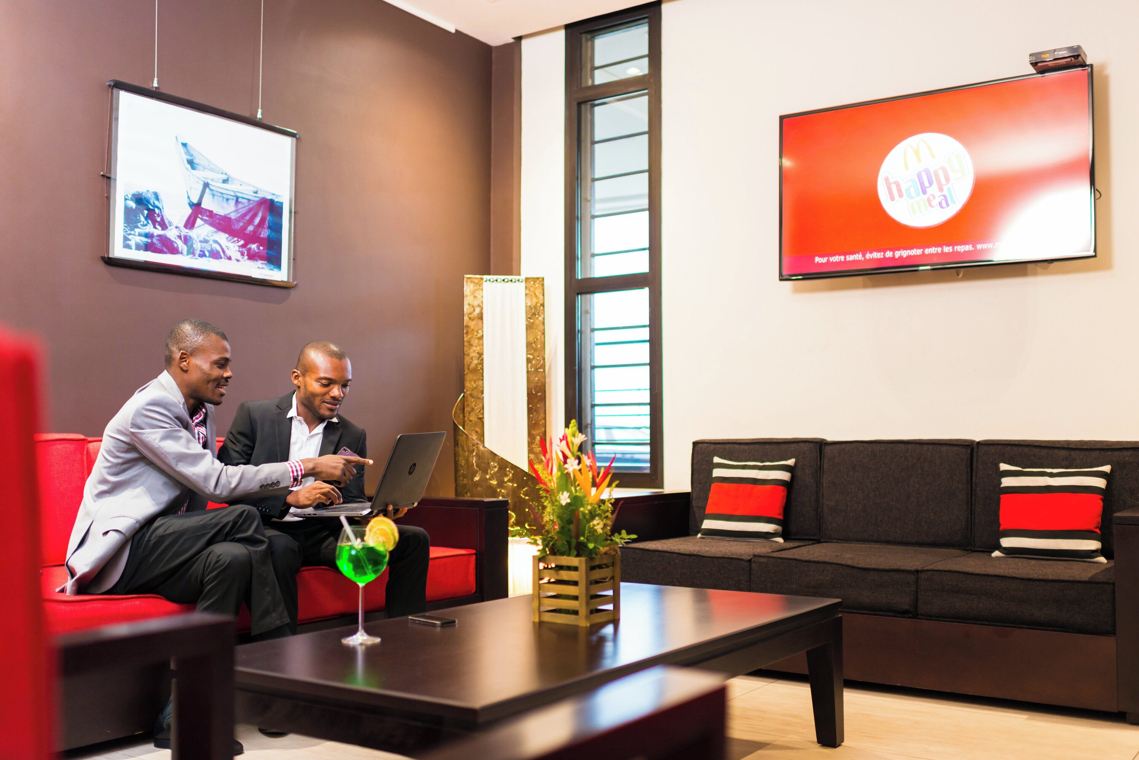 Deux hommes en costume sont assis sur un canapé rouge, devant un ordinateur portable, avec un verre contenant une boisson verte et un vase décoratif rempli de fleurs.