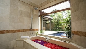 A bathroom with a large bathtub filled with flower petals, a shower, and a view of a pool and greenery outside. - Parigata Villas Resort (Denpasar)