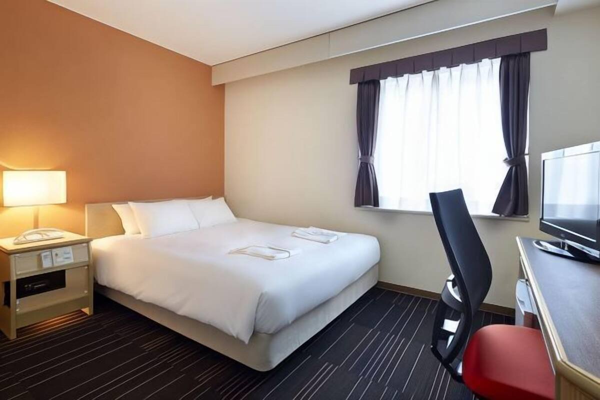 A hotel room with a bed, a desk with a computer, a chair, a lamp, and a window with curtains.