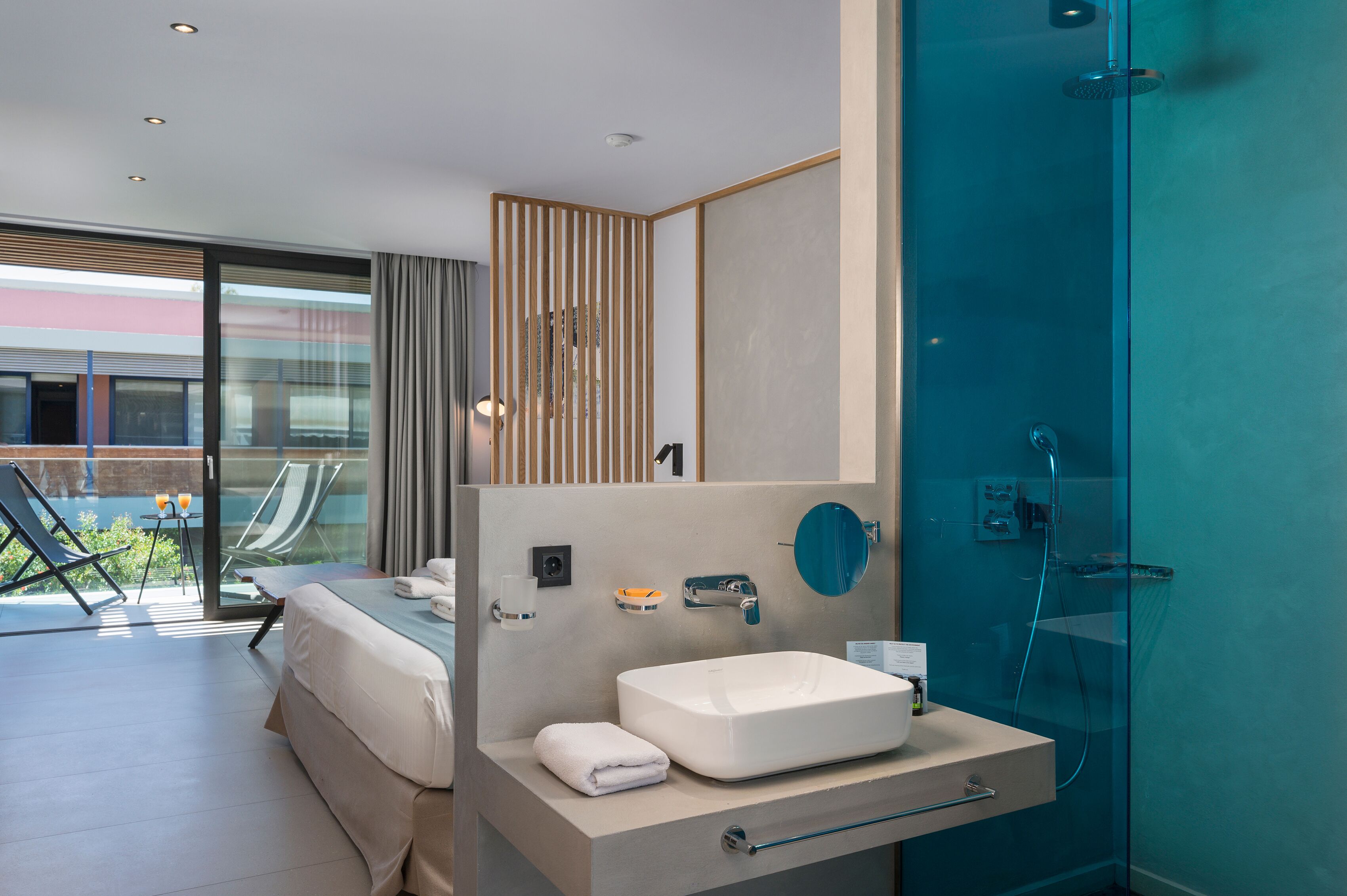 A modern hotel room with a large bed, a bathroom with a glass shower enclosure, and a balcony with a pool and chairs.