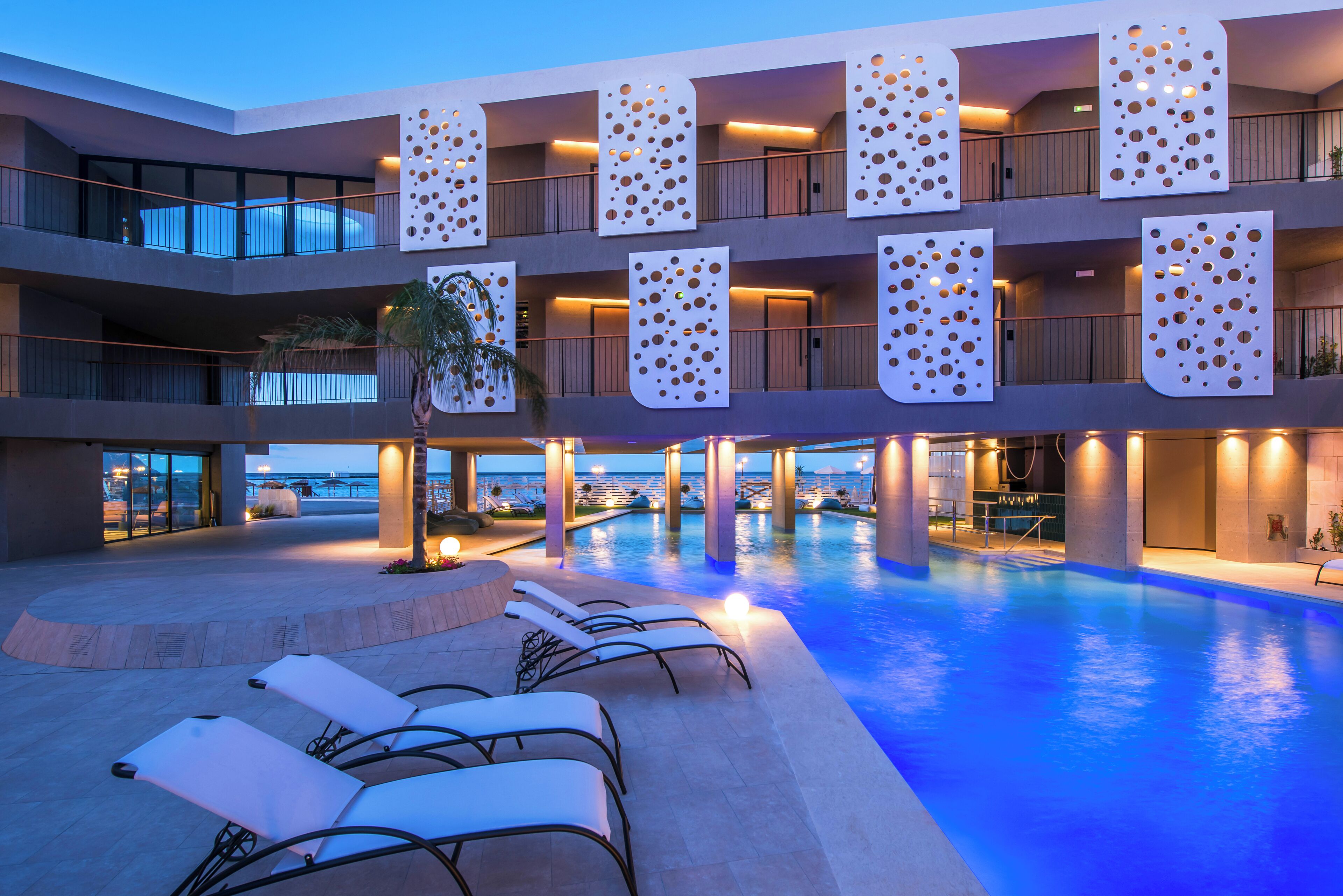A multi-level hotel with a swimming pool, sun loungers, and a view of the sea.