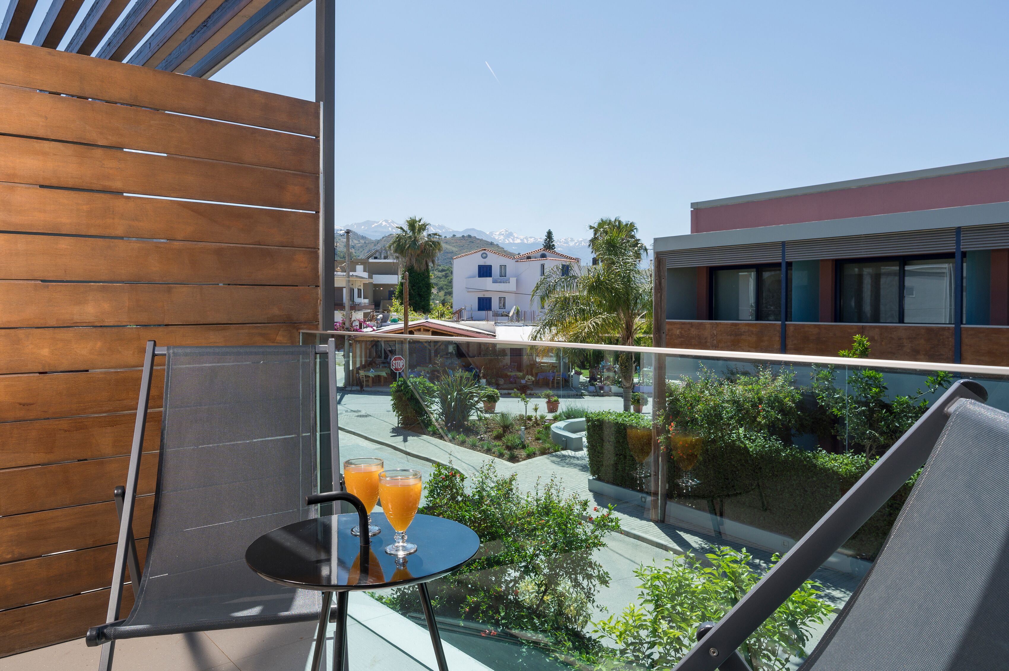 A balcony with a table and two glasses of orange juice, overlooking a courtyard with plants and a mountain in the background.