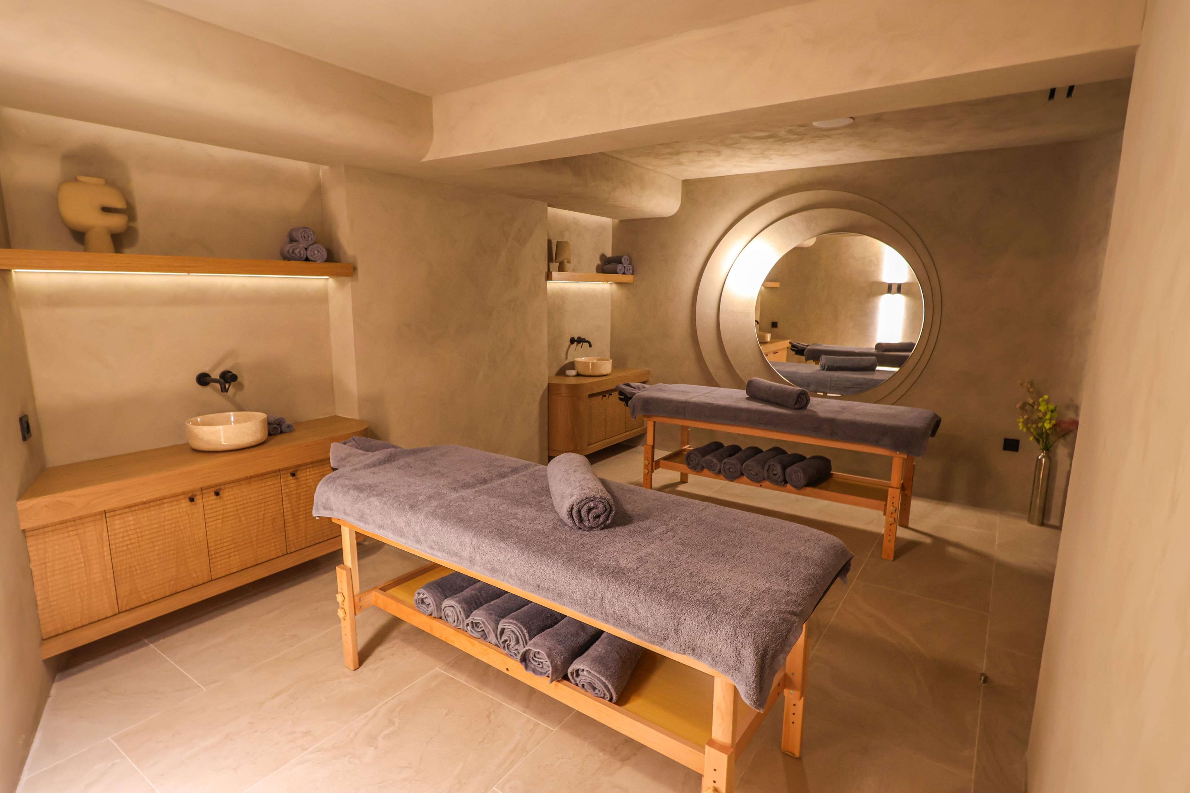 A spa room with a wooden bench, a round mirror, a wooden vanity with a bowl, and a shelf with towels.