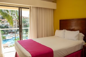 Hotelkamer met een bed, een houten hoofdeinde, een raam met uitzicht op een zwembad en gebouwen, en een gordijn.
