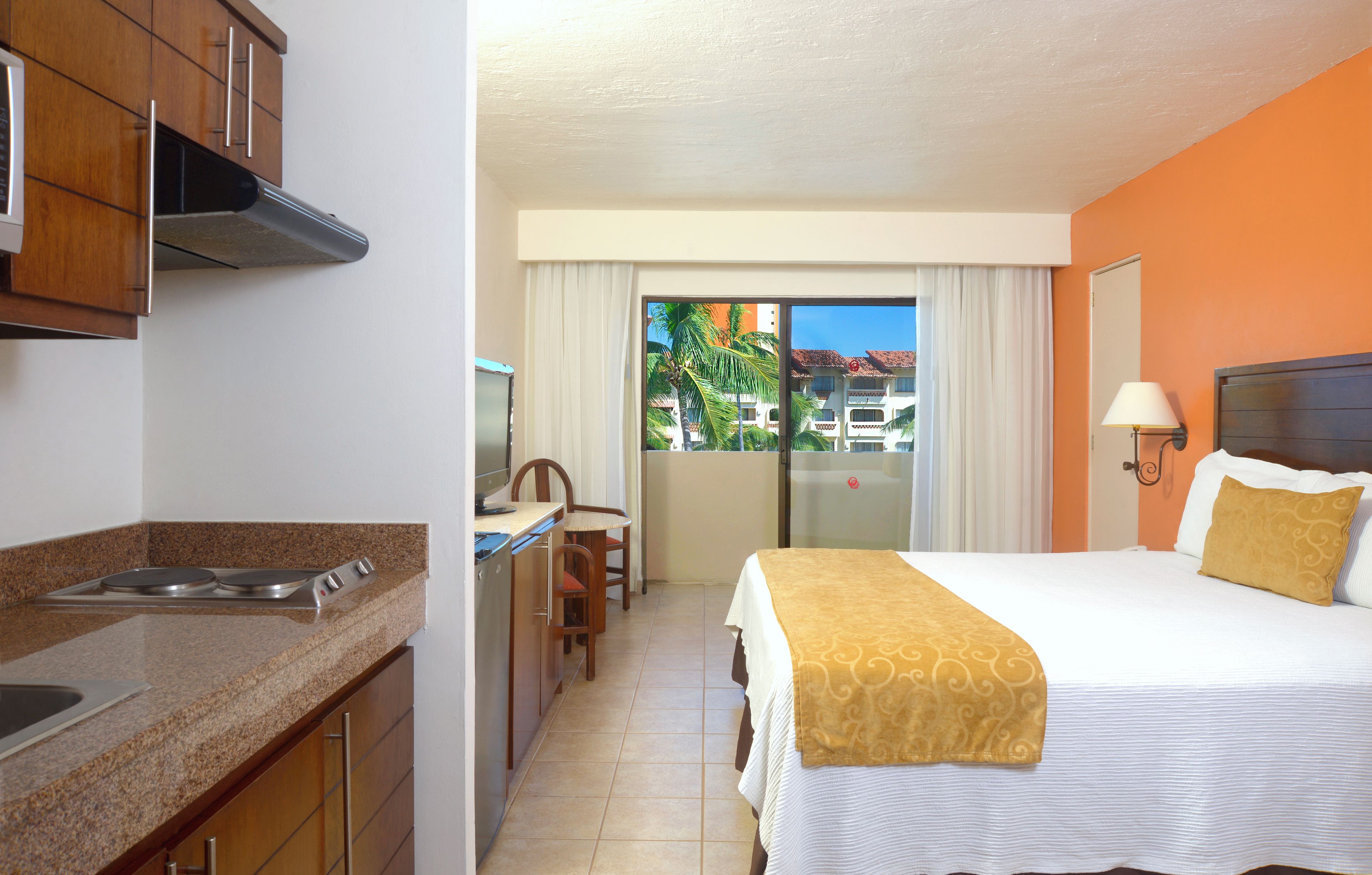 A hotel room with a kitchenette, a bed with a decorative headboard, a dining area with a table and chairs, and a view of a building and palm trees through the window.