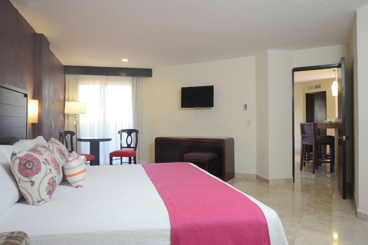 A hotel room with a large bed, a desk, a chair, a TV mounted on the wall, and a door leading to a bathroom.