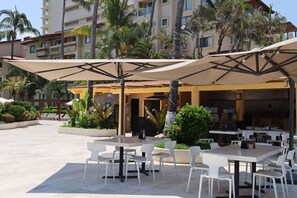 An outdoor dining area with tables and chairs, surrounded by palm trees and a building with a tiled roof. - Canto del Sol Puerto Vallarta All Inclusive (Puerto Vallarta)