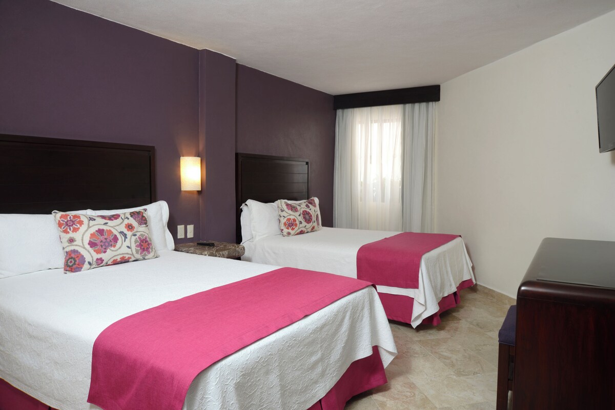 Une chambre d’hôtel avec deux lits, une table de chevet, un téléviseur et une fenêtre avec des rideaux.