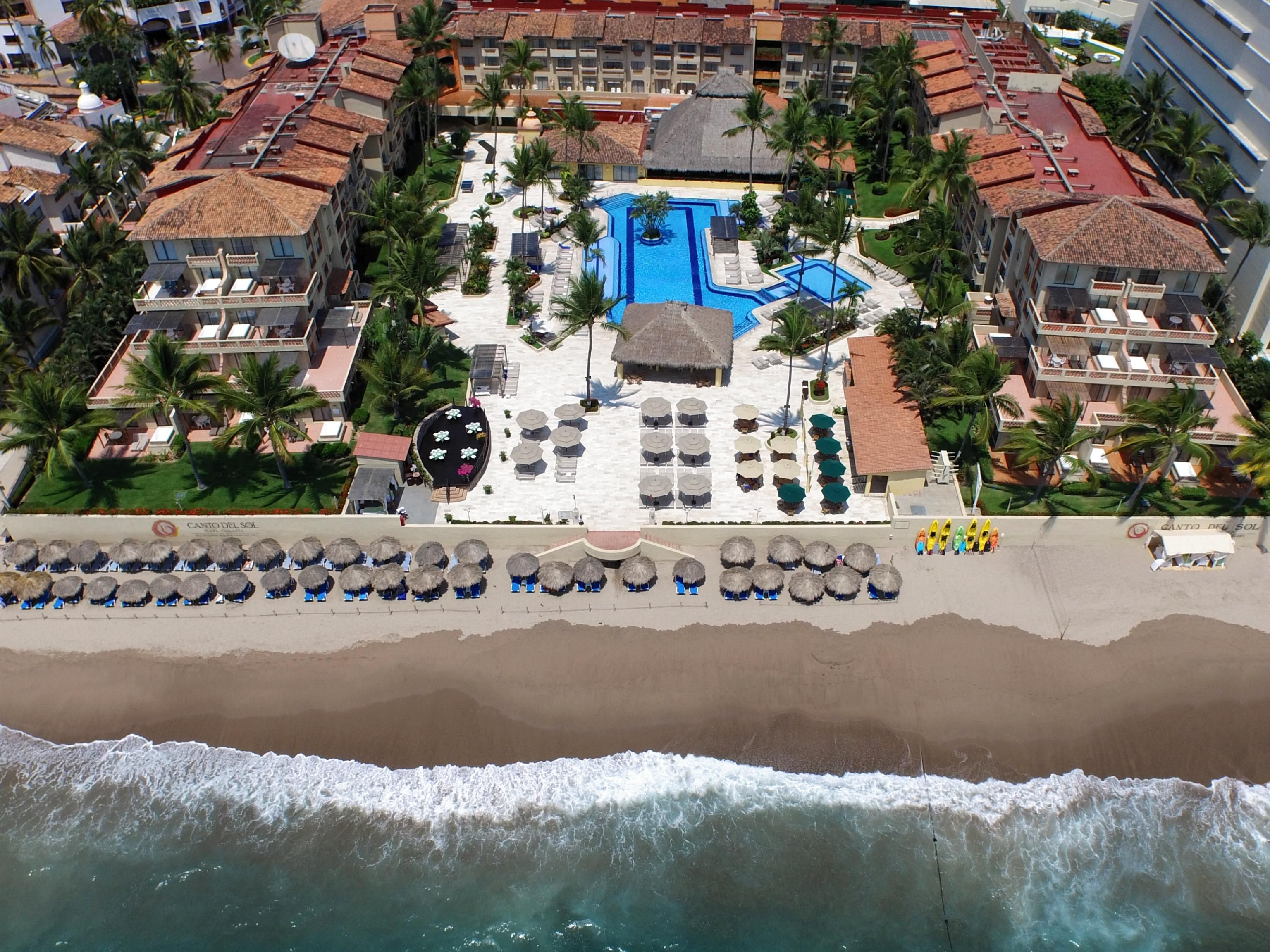 Vista aérea de un complejo turístico con piscina central, varias zonas de descanso y acceso directo a la playa.
