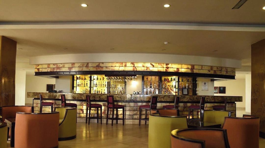 Zona de bar con mostrador curvo, diversas opciones de asientos como taburetes y sillas, y una variedad de botellas y vasos a la vista.