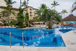 A swimming pool with lounge chairs, palm trees, and a building with balconies in the background. - Canto del Sol Puerto Vallarta All Inclusive (Puerto Vallarta)