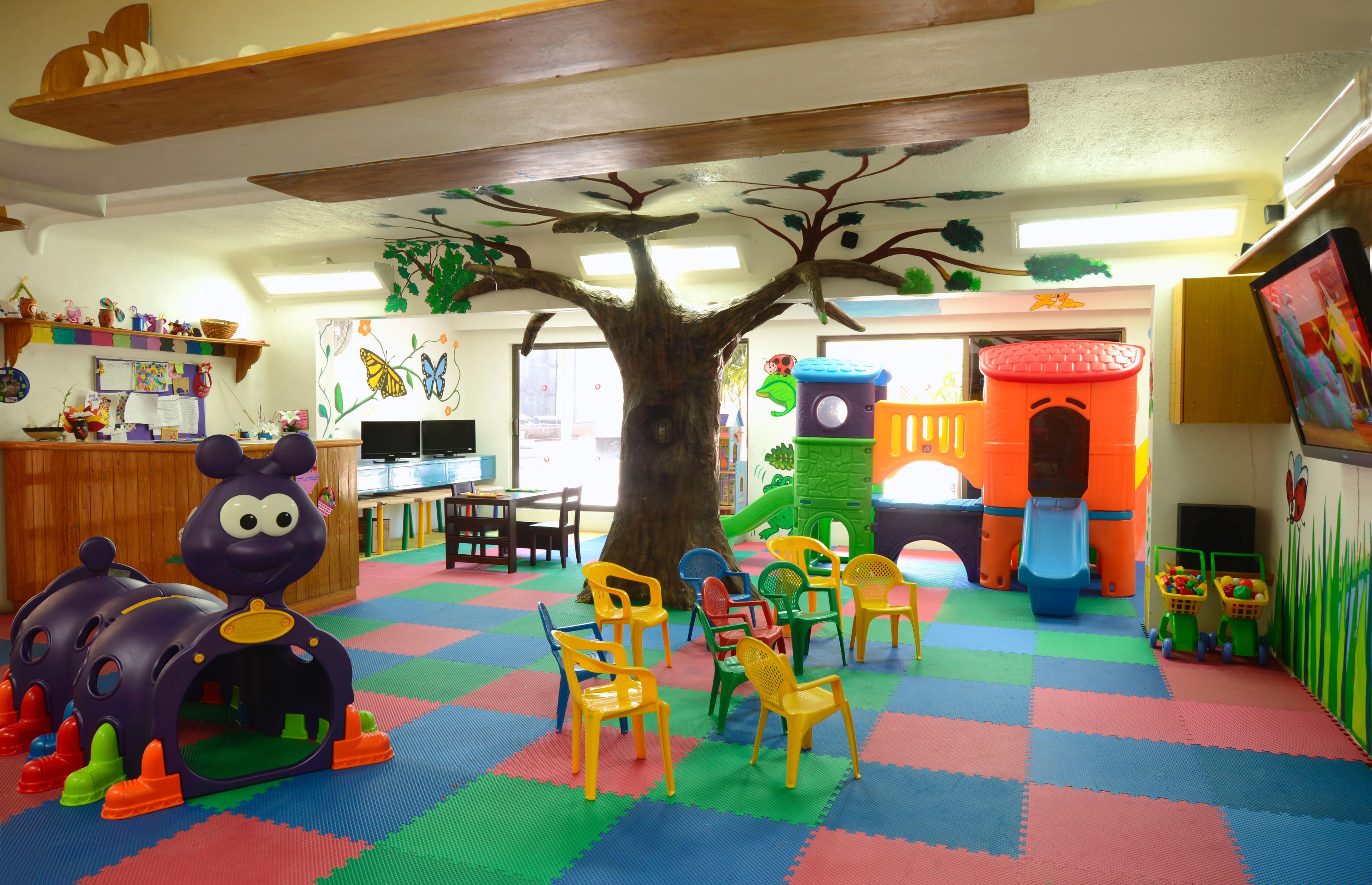 Un área de juegos infantiles con una gran estructura de árbol, alfombras de colores y diverso mobiliario de juego.