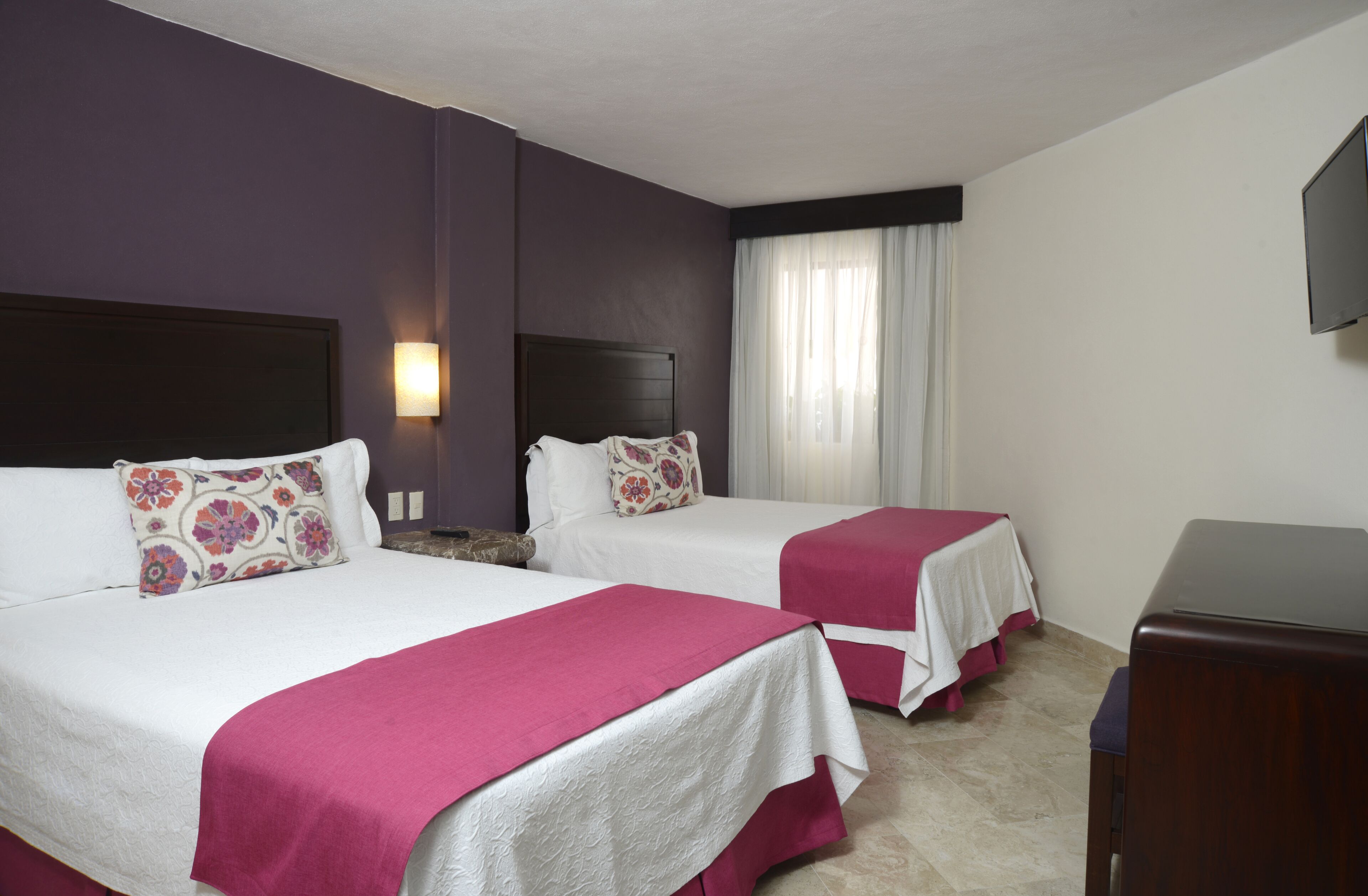 A hotel room with two beds, each with a headboard, a bedside table, and a flat-screen TV mounted on the wall.