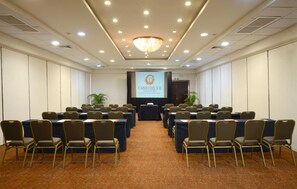 Une salle de conférence avec des rangées de tables et de chaises, un écran de projection et un lustre.