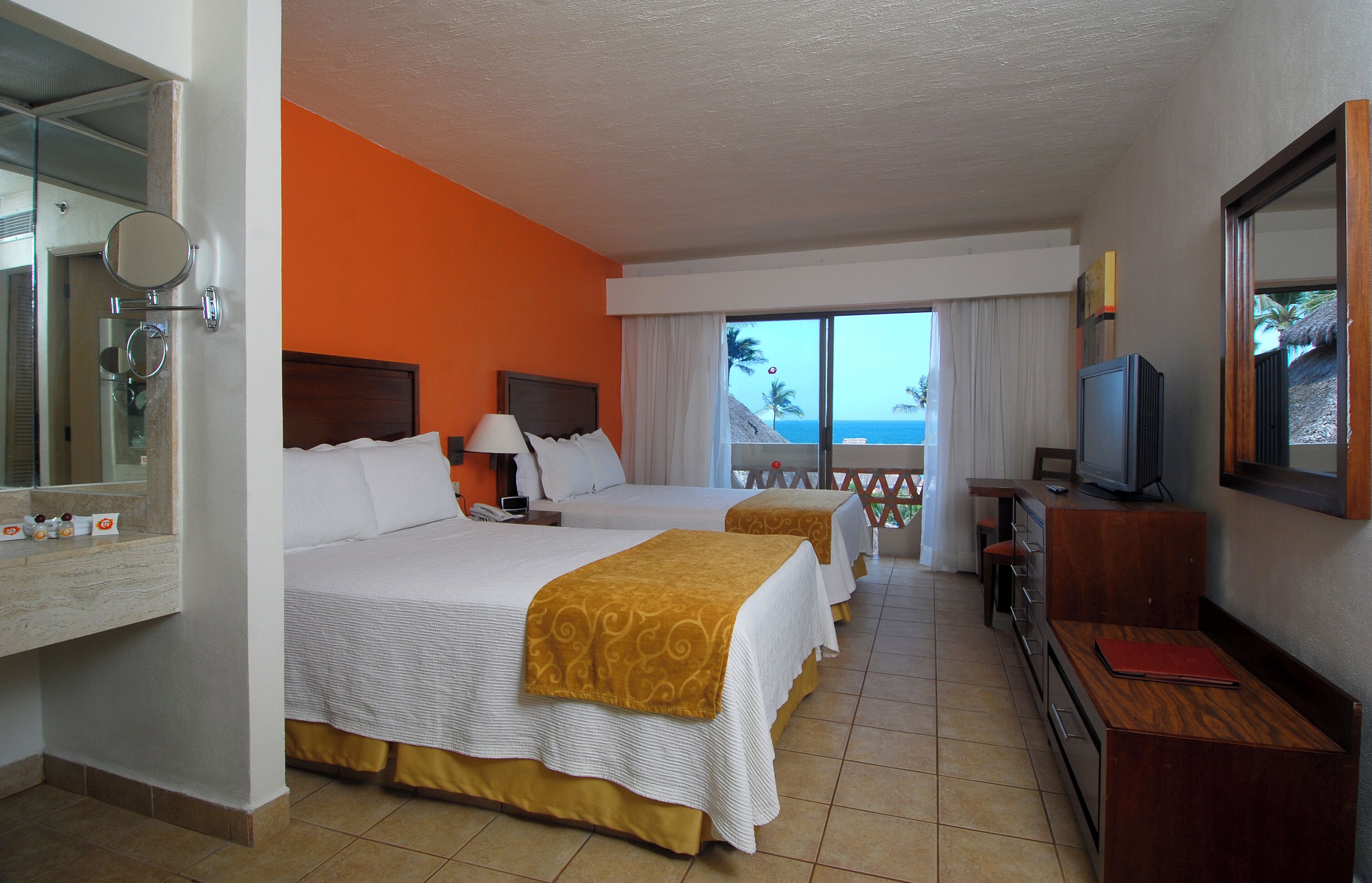 A hotel room with two beds, a TV, a desk, and a balcony with a view of the ocean.